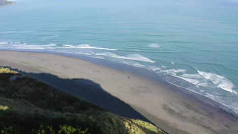 a pair of people ride horseback at a beach in raglan, new zealand along hillsides and mountains taken by drone