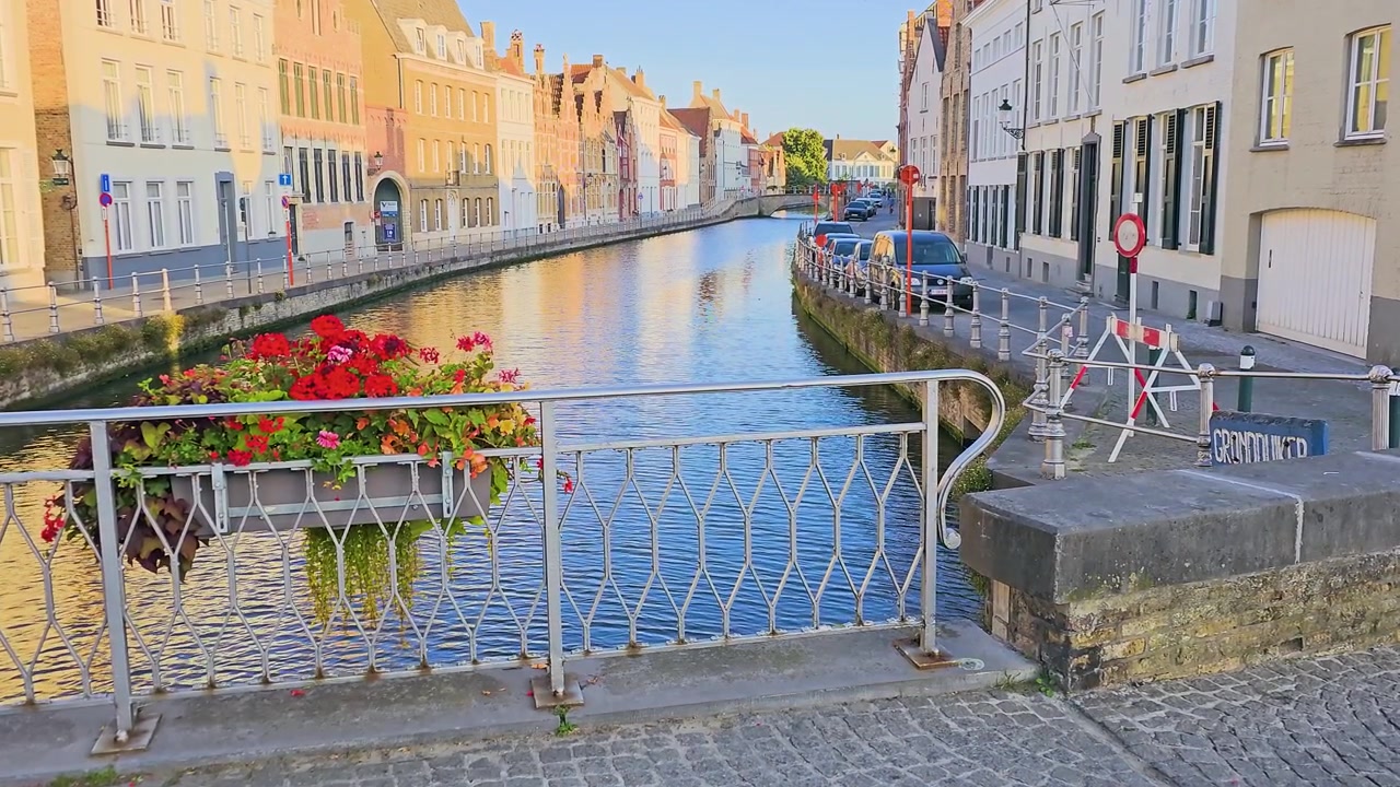 Scena di una storica città con canali, con fioriera sulla ringhiera del ponte, acqua calma, vecchi edifici in mattoni, auto parcheggiate e calda luce del tramonto, un affascinante sfondo di viaggio europeo.