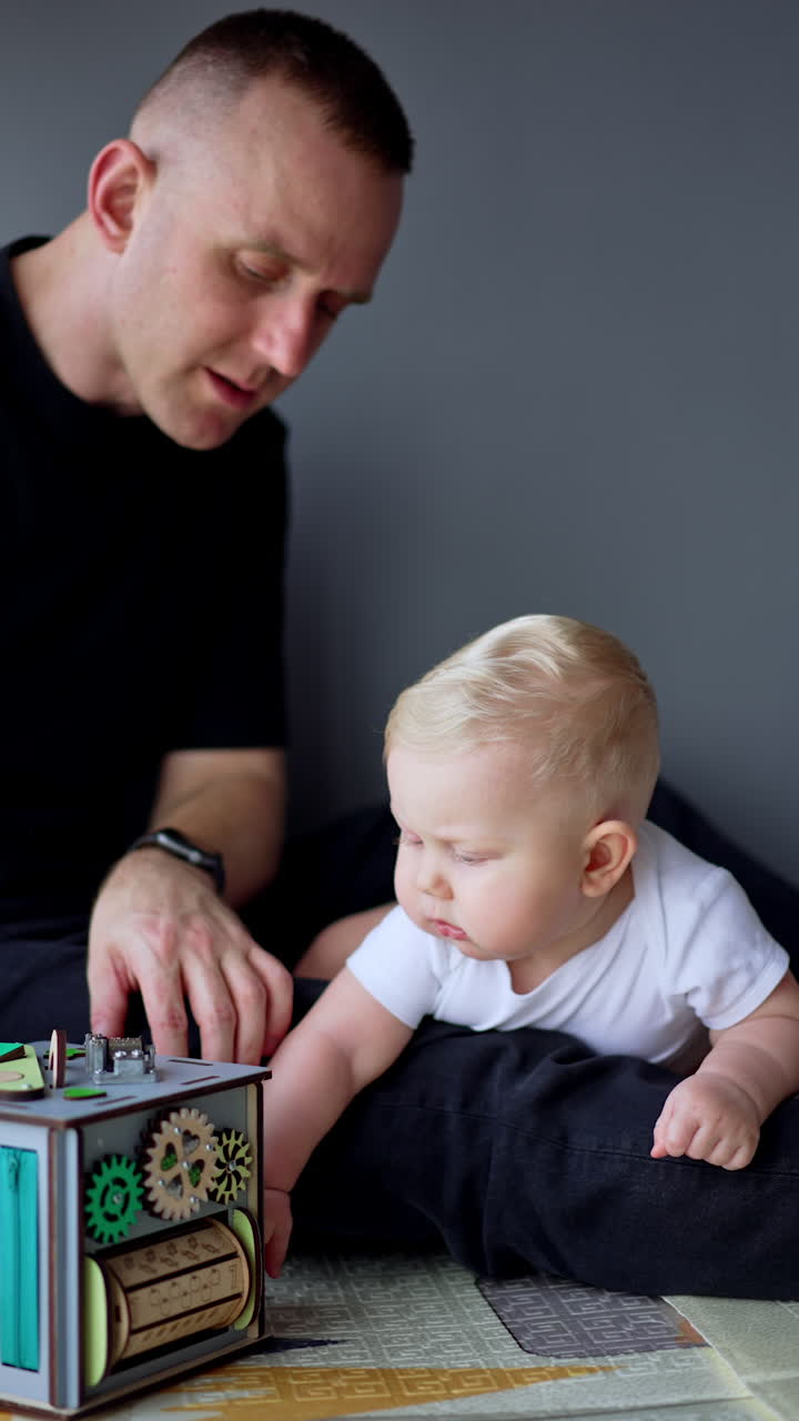 Caucasian father showing the toy to his son talking to him. Lovely blond baby boy looking attentively. Vertical video.