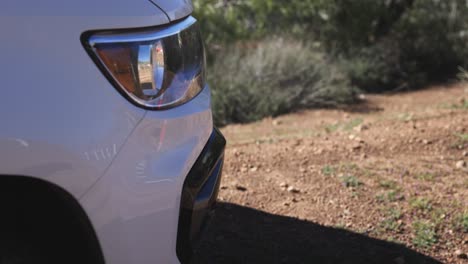 static shot of the headlight and front end of a white car on a dirt road with vegtetation in the background