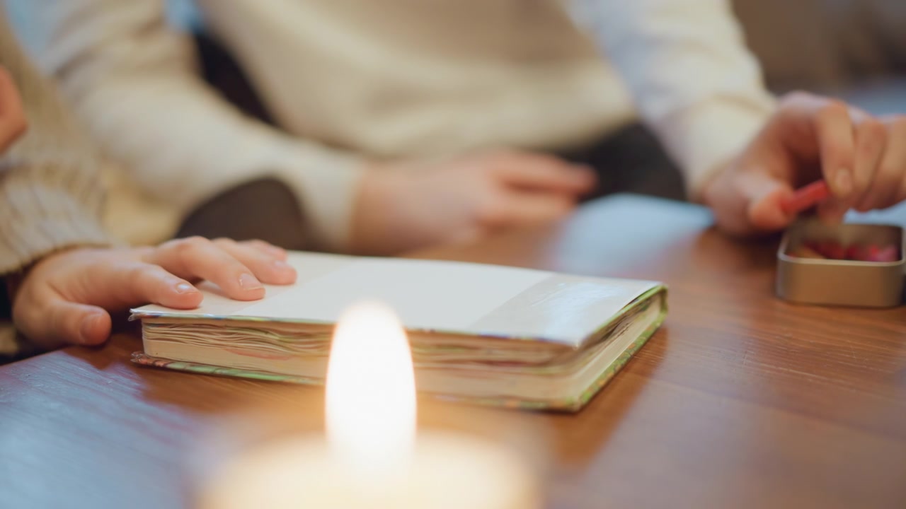 Primo piano di candela accesa su tavolo di legno con quaderno e mani sullo sfondo. Le pagine del quaderno sono impilate, mostrando bordi colorati sotto una luce soffusa. Le mani riposano vicino al libro mentre un'altra mano