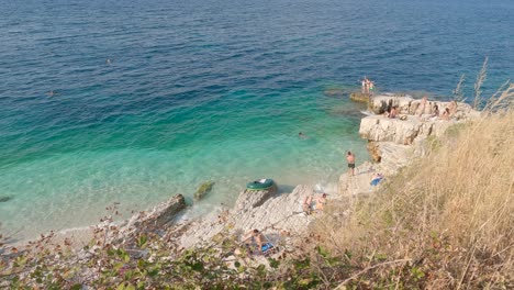 tourists in turquoise rocky beach in kassiopi coastline, panning shot from above, corfu island