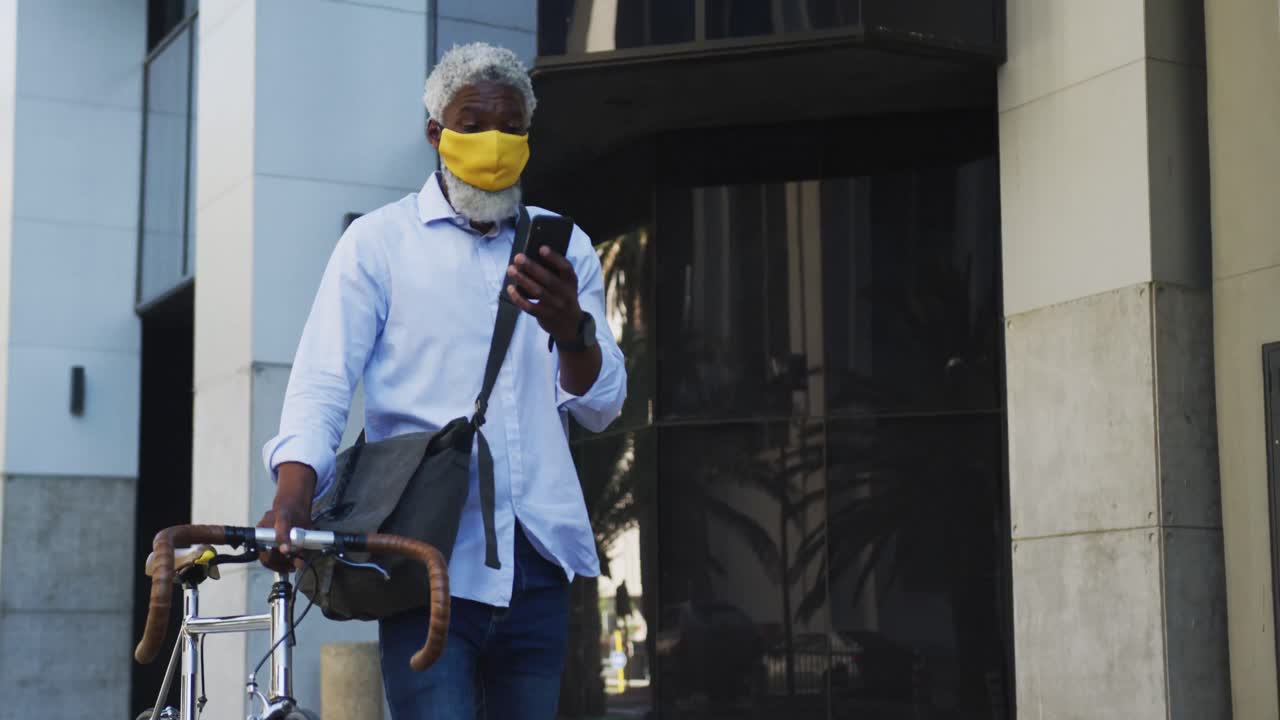 African american senior man wearing face mask with bicycle using smartphone in corporate park