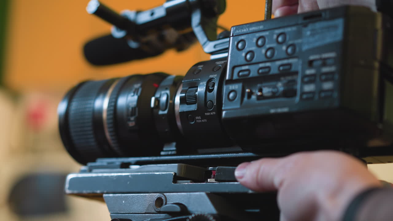 Close-up of camera lens mounted on tripod quick release plate. Focus on camera controls and zoom lens. Background features blurred orange wall. Hand adjusting camera setup