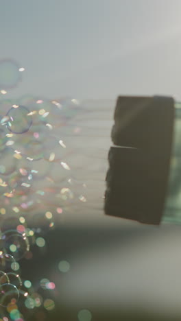 close-up of a plastic bubble gun releasing a stream of colorful bubbles under a bright, slightly cloudy sky. the bubbles sparkle as they float through the air