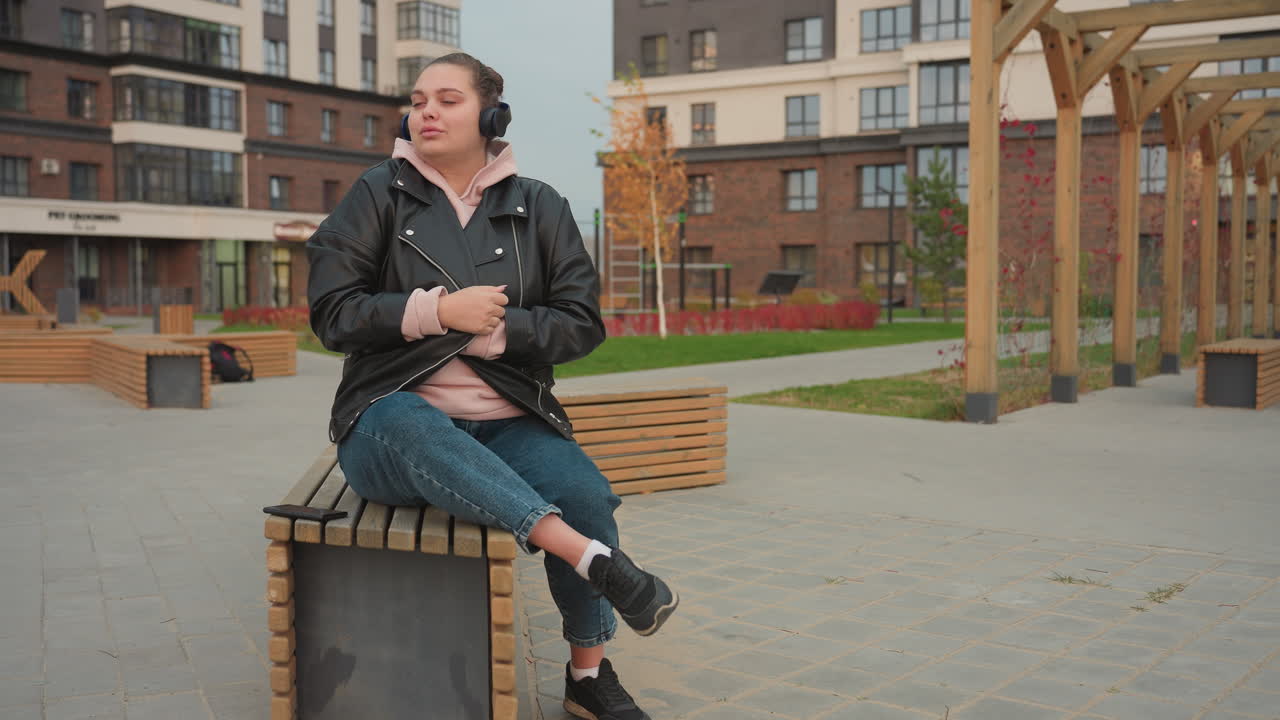Elegant woman seated on wooden bench in stylish black jacket and pink hoodie adjusts jacket while enjoying music through headset in open space surrounded by cool evening breeze