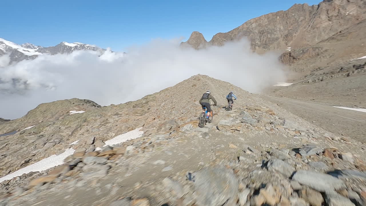 FPV drone follows two mountain bikers descending steep, exposed alpine trails above Saas-Grund and Saas-Fee on a clear bluebird summer day, showcasing epic terrain and speed