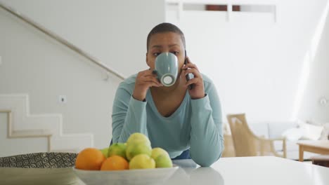 Happy-african-american-plus-size-woman-drinking-coffee,-talking-on-smartphone-in-kitchen