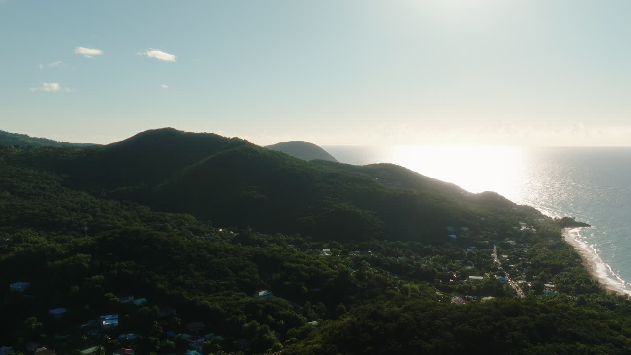 Drone moves laterally and descends along the green hills of Guadeloupe toward the sea, with sunlight reflecting on the water