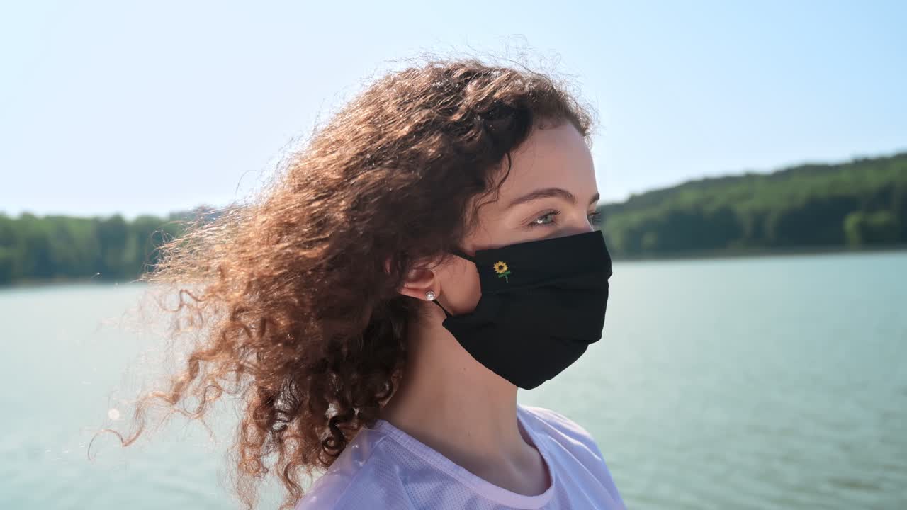 Young curly woman looking at the lake while wearing a black protective mask. Blowing by the wind in a sunny day. Close up. Corona Virus idea