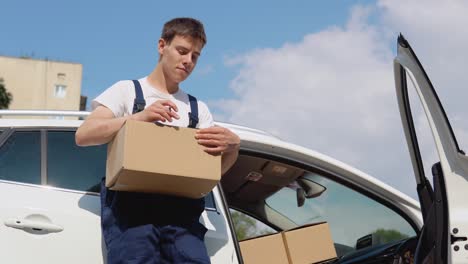 movimiento y entrega de bienes fabricados. el conductor llena los documentos de carga y mira a la cámara. un empleado sostiene una caja en sus manos y se para junto a un coche cargado de cajas