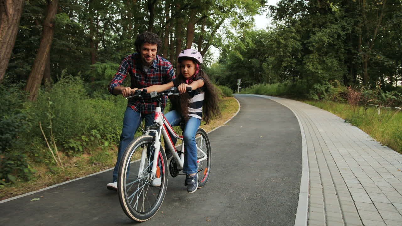 클로즈업: 어린 소녀의 초상화. 아빠가 자전거를 타는 법을 가르치고 있다. 그녀를 놓아주고 있다. 웃고 있다. 카메라를 움직이고 있다. 흐릿한 배경.