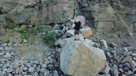 a man in a black t-shirt and a backpack comes to the edge of a large stone and raises his hands up. tourist got to the center of stone quarry.