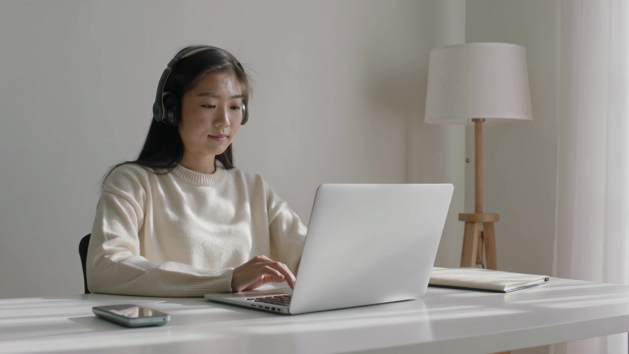 自宅の机で、クリーム色のセーターを着てヘッドホンを着用したアジア人女性が、音声を聞きながらノートパソコンでタイピング作業をしている。