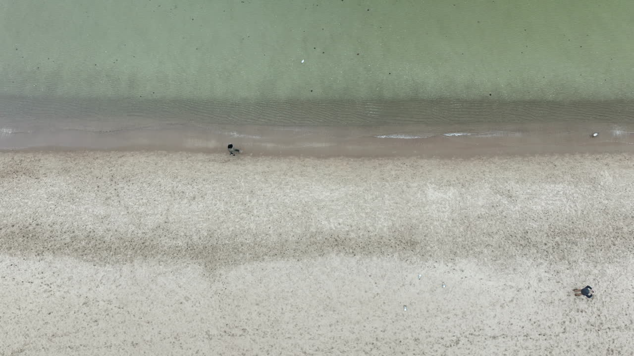 Overhead drone shot of sandy shoreline with two people walking along the water leaving footprints
