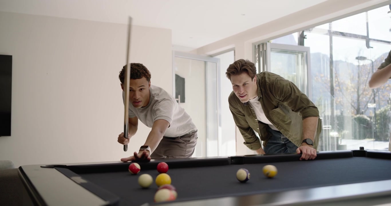 Diverse men after cue swap watching African-American man taking shots on black pool table for fun