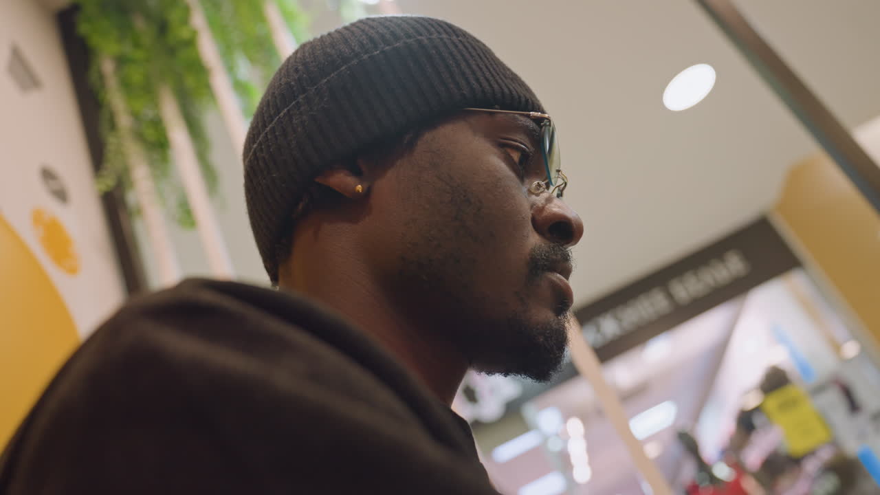 Black man in beanie focused gaze, side profile in retail lounge checking tablet and notes, determined entrepreneur energy, soft overhead lights and cinematic color grading, candid productivity scene