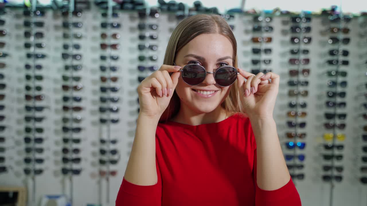 Portrait of attractive girl choosing sunglasses. Female buyer is posing on camera in new trendy glasses in the optics store.