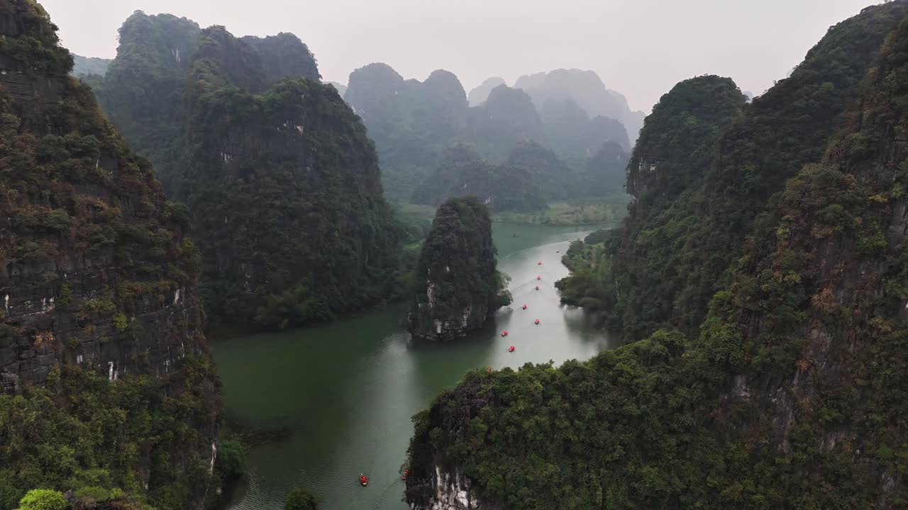 Aerial view of Trang An in Ninh Binh, Vietnam, showing limestone karst mountains, a winding river, lush greenery, and red boats navigating through the scenic landscape