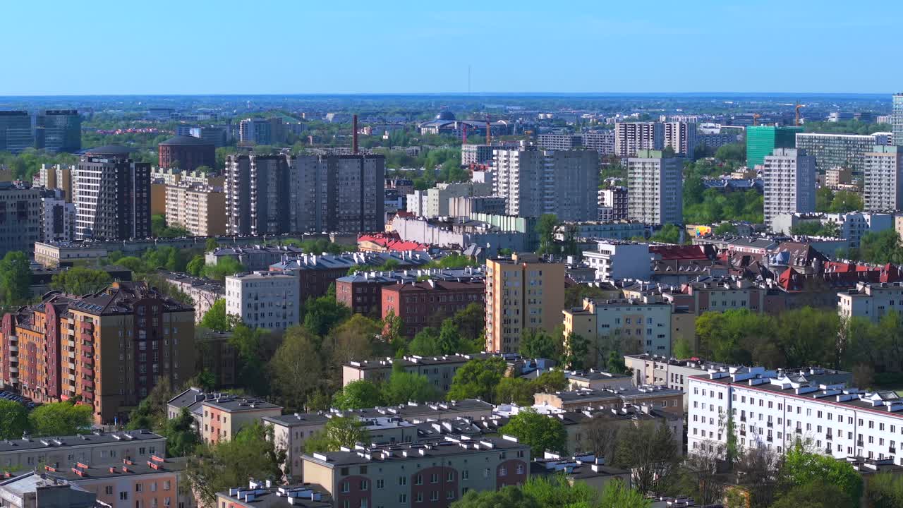 Aerial establishing shot of Warsaw’s Wola district showing dense urban skyline, mid-rise apartments, and green pockets under a clear sky