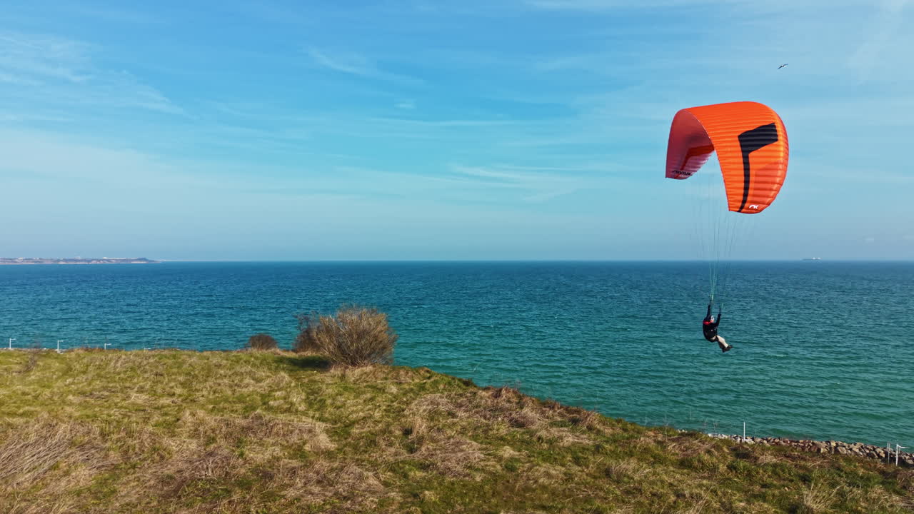 Paraglider soars above green hill near ocean on clear day with clear sky and distant city in background