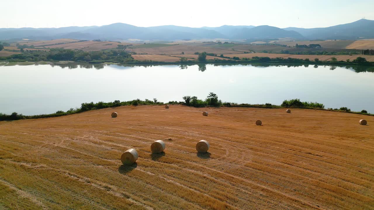 Golden hay bales dot the tuscan countryside near a tranquil lake, reflecting the sky and surrounding hills in its clear waters, creating a peaceful and picturesque scene