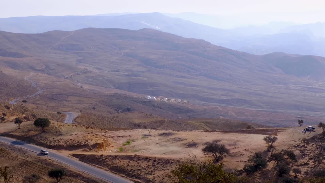 carro branco dirigindo através de uma paisagem remota e montanhosa do deserto com acomodação de tenda de bolha à distância na jordânia, oriente médio