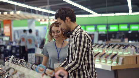 In-the-electronics-store-female-consultant-gives-professional-advice-to-a-young-man.-He-considers-to-buy-a-new-smartphone-and-needs-expert-opinion.-Store-is-modern,-bright-and-has-all-the-new-devices.-Blurred-background.-Slow-motion