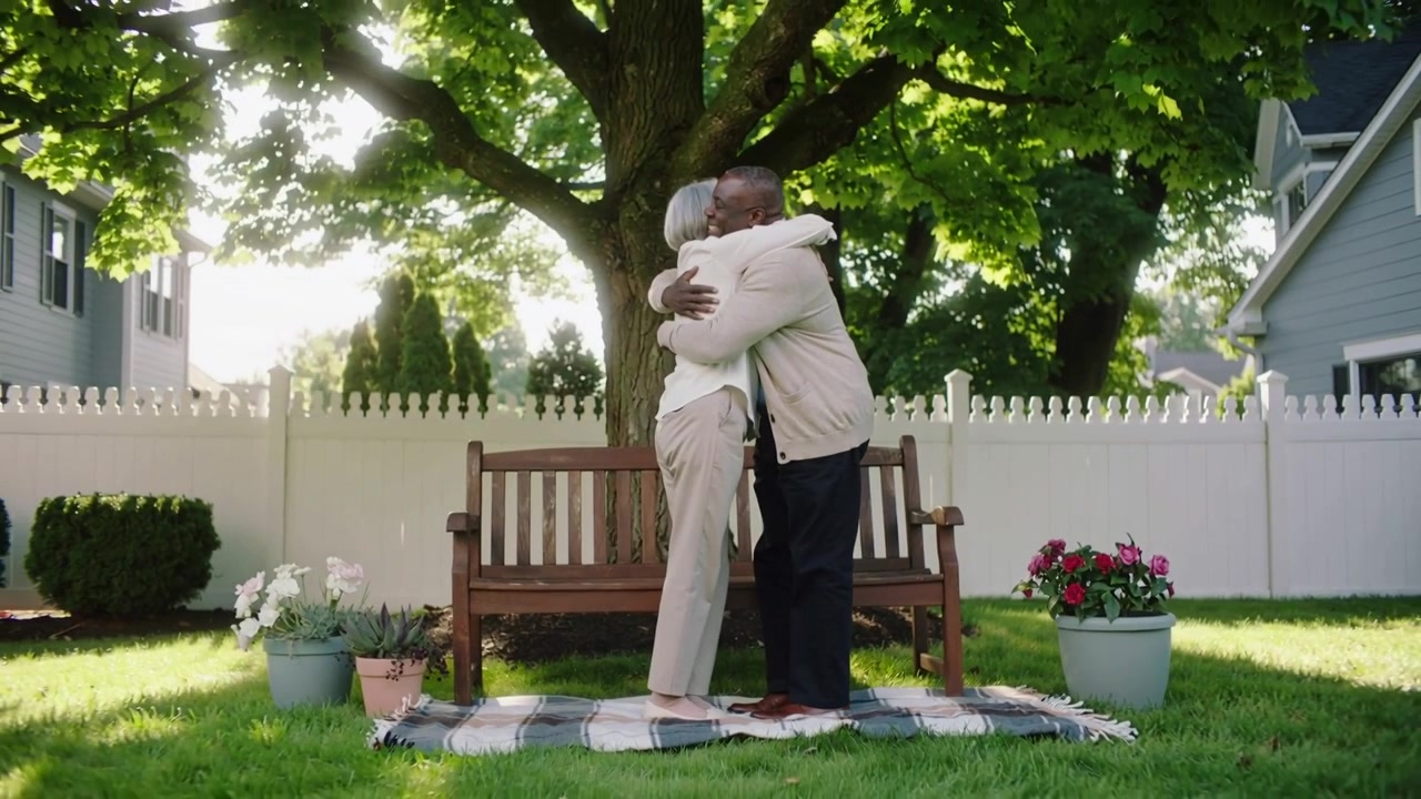 Kissing senior couple rising, holding and standing on bench in yard with plaid blanket and flowers