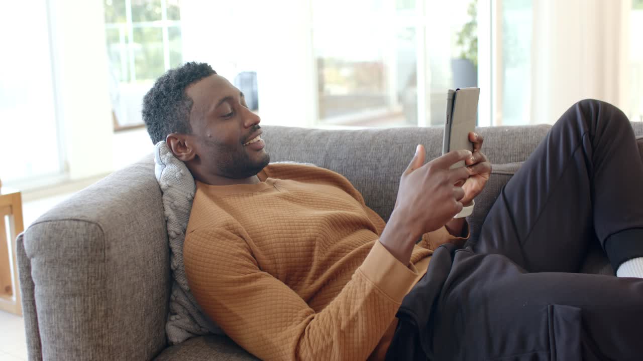 African American man lifting tablet, reading content while reclining on gray sofa with pillow