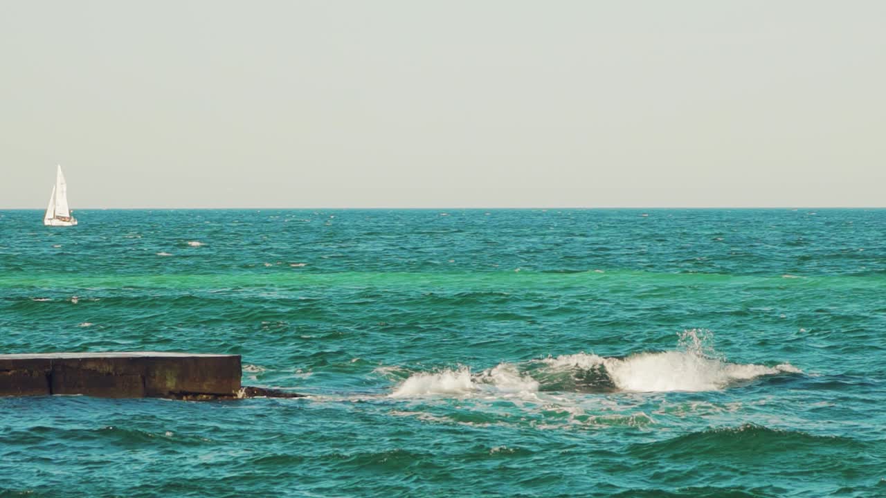 a white sailboat is floating in the sea near the coast on the background of an old pier in the summer. Seascape. Slow motion