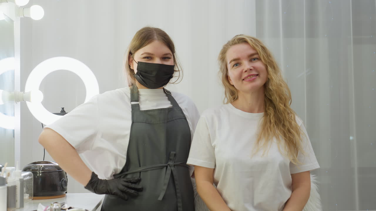 Client and beautician pose side by side in salon, both looking at camera with relaxed expressions, beautician wearing gloves and apron, standing confidently beside seated client near mirror and tools