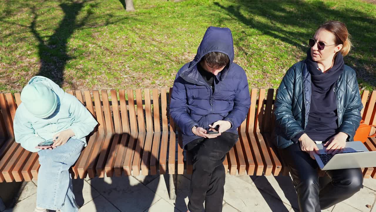 Ihmisiä istumassa puiston penkillä uppoutuneina älypuhelimiinsa ja kannettavaan tietokoneeseensa