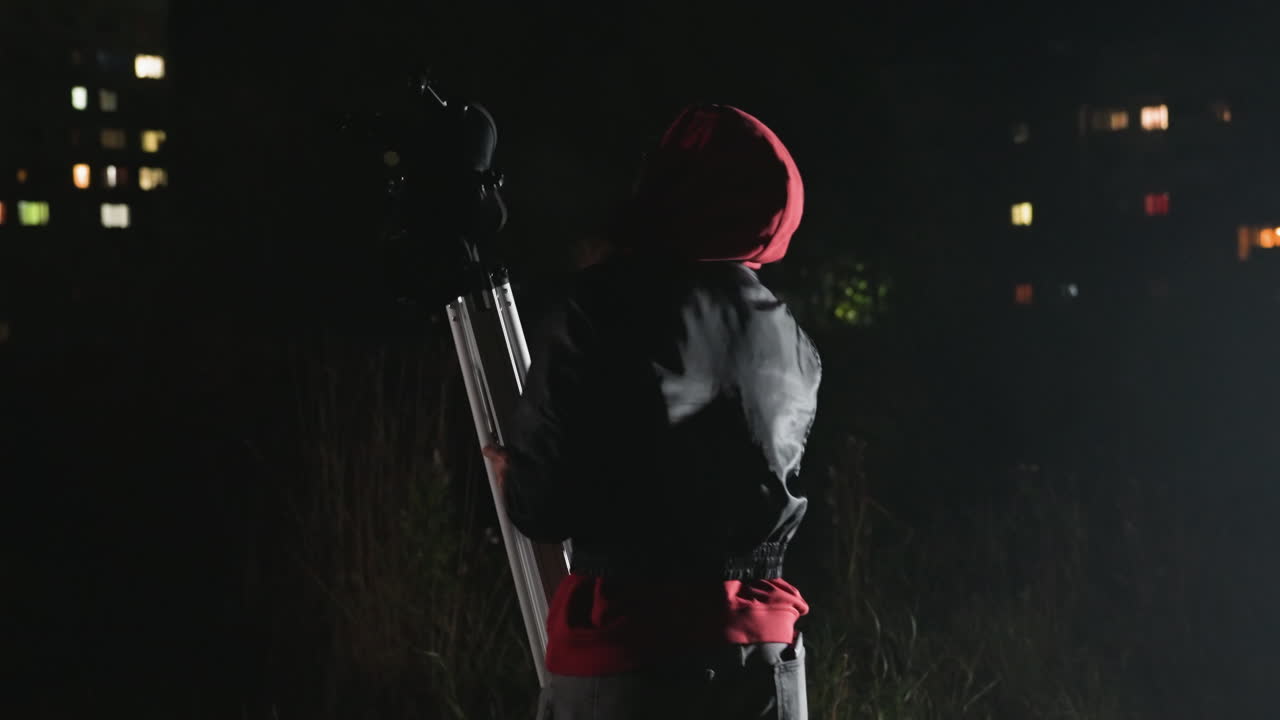 Silhouette notturna con telescopio vicino ad alcuni appartamenti, un osservatore incappucciato sistema un treppiede in un campo ombreggiato, regola l'oculare verso il cielo scuro, le luci delle finestre della città brillano sullo sfondo, atmosfera tesa e riflessiva