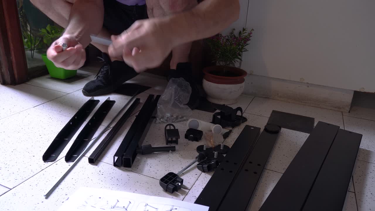 Man sitting on the floor assembling furniture while holding an instruction manual, surrounded by tools, metal parts, and hardware components in a home environment