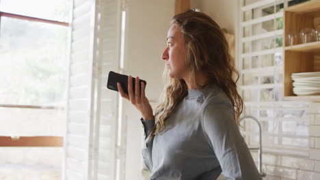 Nachdenkliche-Kaukasische-Frau-In-Der-Sonnigen-Landhausküche,-Die-Ihr-Smartphone-In-Der-Hand-Hält-Und-Aus-Dem-Fenster-Schaut