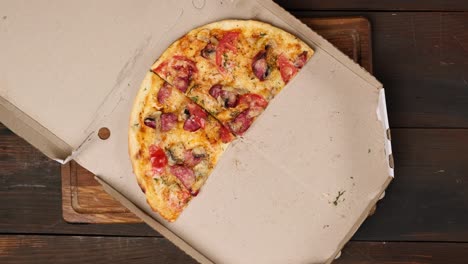 baked round pizza with cheese, tomatoes, sausage and mushrooms cut into pieces in a white cardboard box, female hand takes a slice