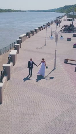 happy young couple walks joining hands along modern waterfront near calm river on sunny day bird eye view