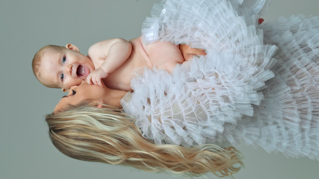 Happy smiling baby boy held by his loving mom. Woman kisses her child on the cheek. Vertical screen.