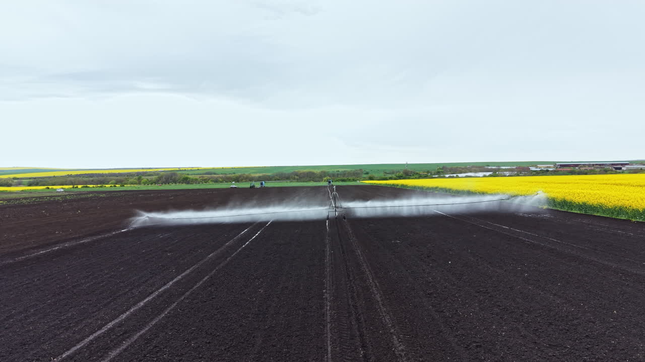 Attrezzatura agricola irrora liquido su terreno scuro in un campo accanto a fiori giallo brillante durante una giornata nuvolosa in campagna.