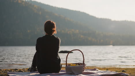 mujer disfruta de la vista panorámica en el picnic. relajarse en la orilla con vistas a las montañas onduladas con densos bosques. dama solitaria admirando la belleza de la naturaleza sentada en cuadros con canasta de comida