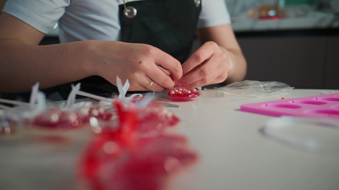 Una persona assembla con cura cuori di caramelle rosse su un tavolo pulito. Le mani si concentrano intensamente sulla lavorazione dettagliata. Stampi rosa brillante e caramelle incartate circondano l'area di lavoro