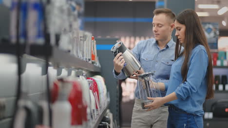 the couple in casual clothes in the electronics store chooses an electric kettle for their kitchen, holding in their hands and reading and reading the price tags with the characteristics