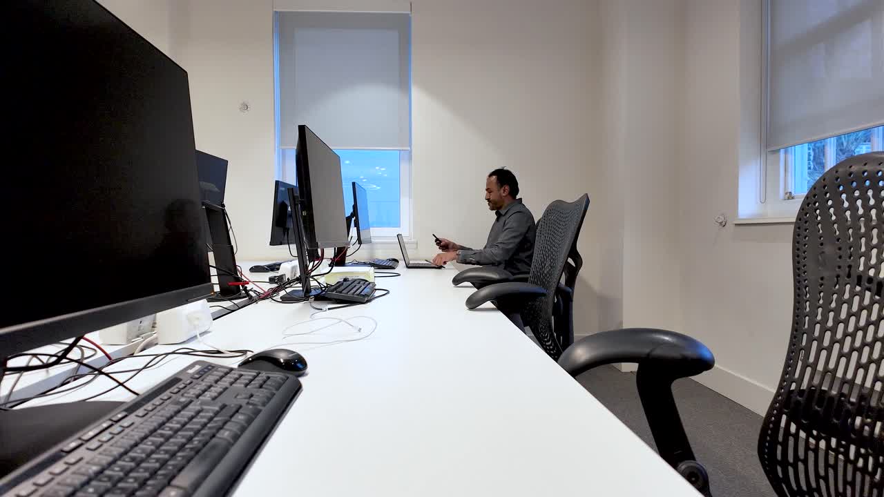 A South Asian man arranges equipment at an empty office desk with multiple monitors and ergonomic chairs