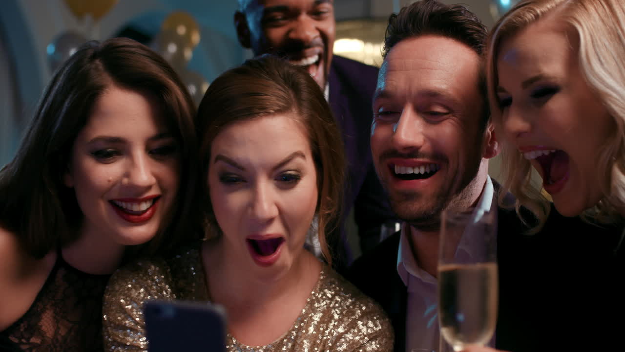 섹시한 친구들이 재미있는 바이러스 비디오를 보고 웃고 화려한 패션 파티에서 스마트폰을 보고 있습니다.