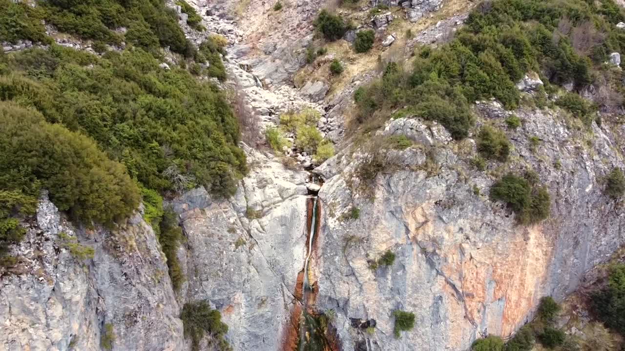 Ripresa cinematografica con drone in allontanamento che rivela una stretta cascata di montagna che scorre lungo ripide scogliere rocciose. Vista dall'alto del paesaggio selvaggio e aspro e del flusso d'acqua naturale. Nessun audio incluso