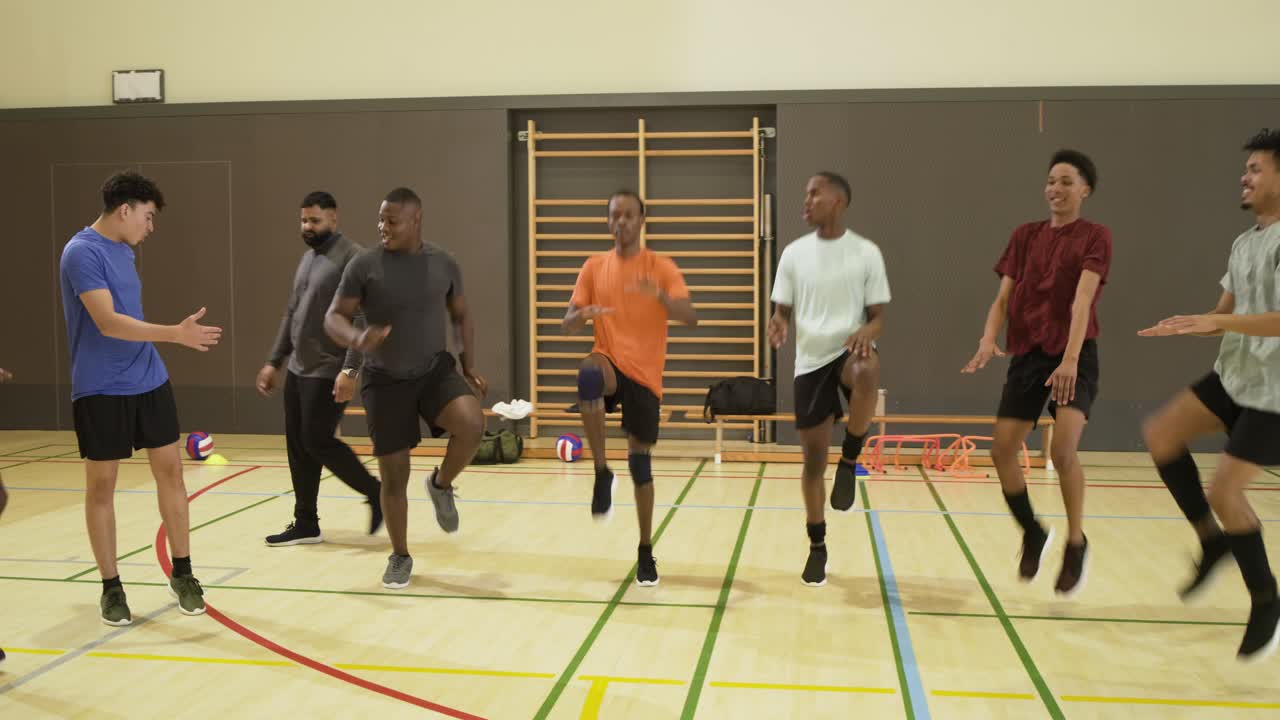 Mandlige atleter varmer op med høje knæløft i fitnesscenter efter anvisninger fra en træner med grå kasket ved nettet
