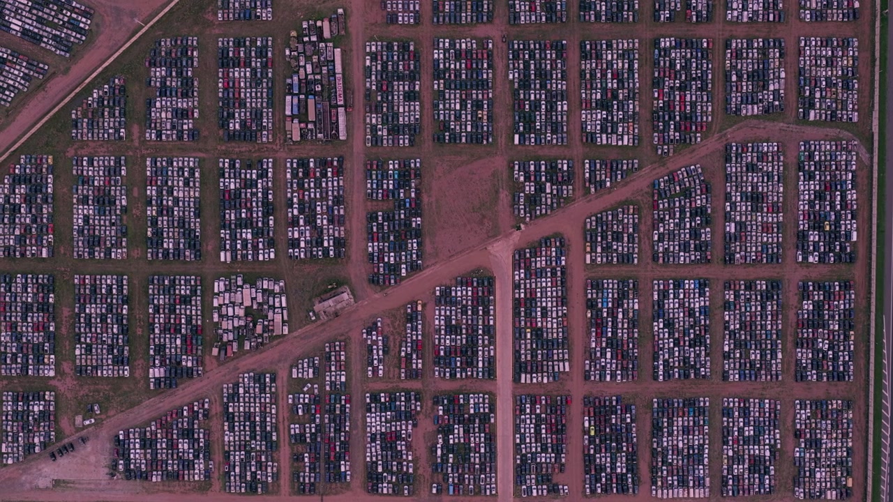 Zenithal aerial pan left across thousands of vehicles organized in dense rows at a large automotive storage facility beside a highway.