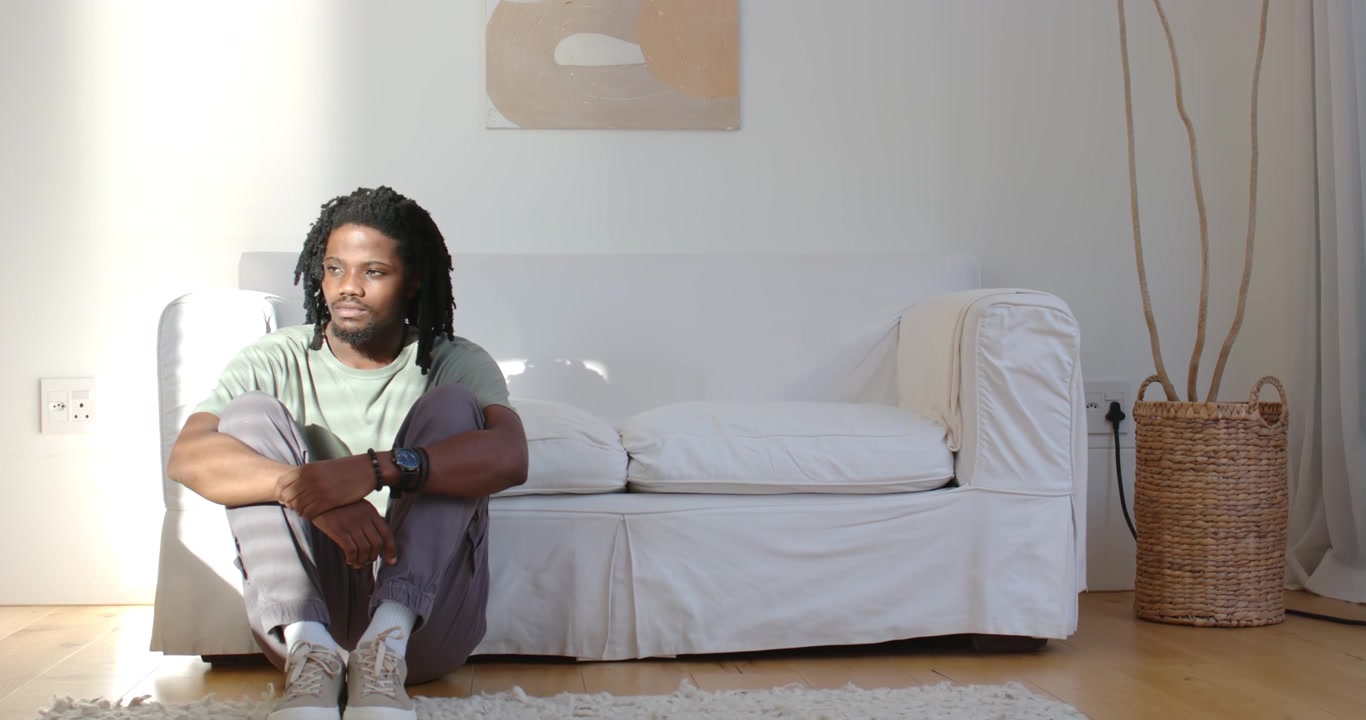 Uomo afroamericano seduto sul pavimento accanto al divano, che abbraccia le ginocchia mentre la luce del sole si sposta, spazio per il testo.