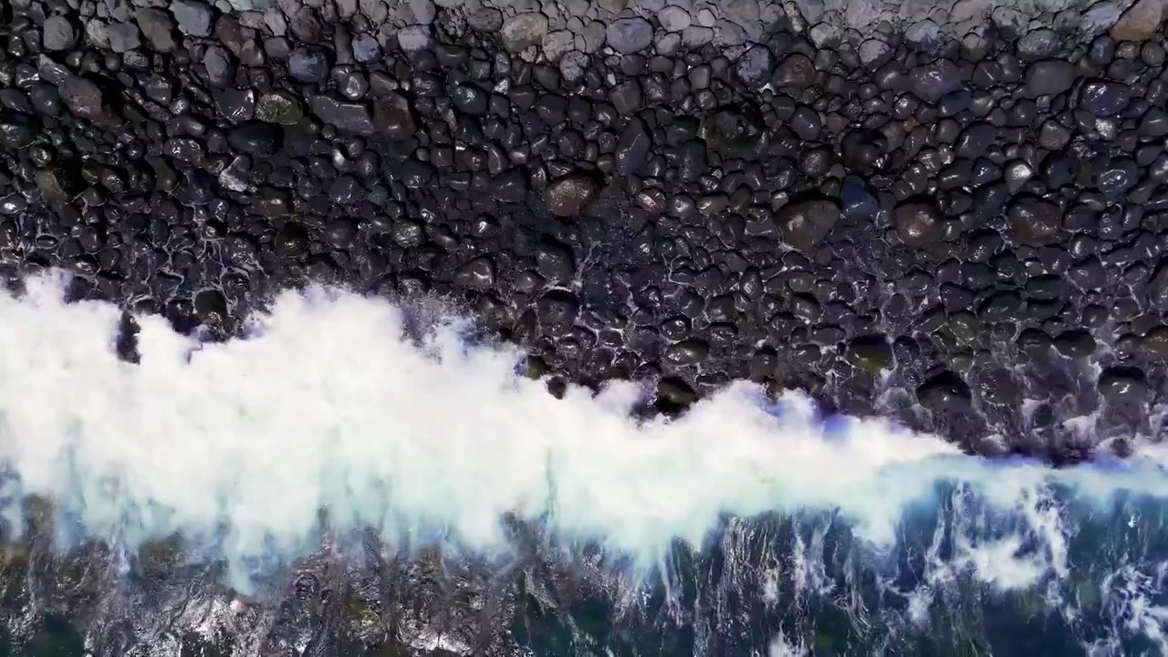 Aerial view of waves crashing on a rocky shore, clear water and wet stones visible